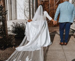 Bride's flowing veil trails behind as couple walks hand in hand along pathway.
