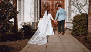 Couple walking towards wooden church doors with flowing white wedding dress train on concrete path.