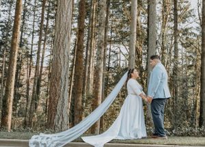 Couple in wedding attire standing among tall pine trees with long flowing veil.