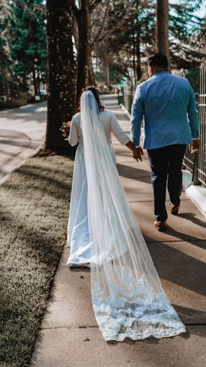 Long lace wedding veil trails behind couple walking on tree-lined pathway.