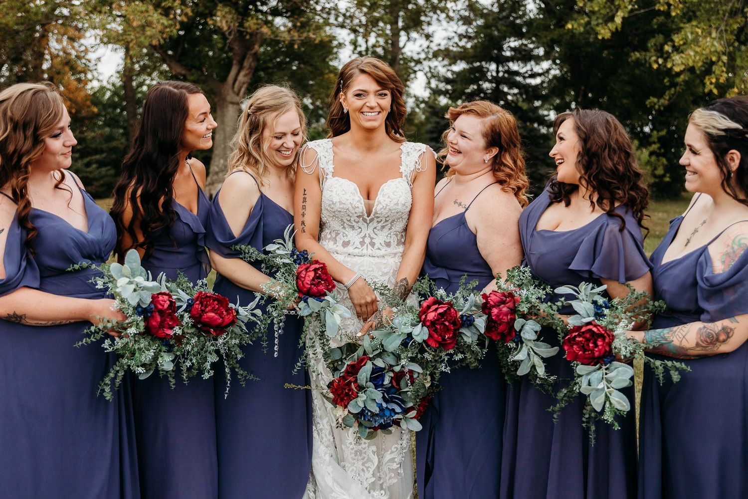 Bridesmaids in navy blue dresses gather around bride while holding vibrant burgundy and green floral bouquets.