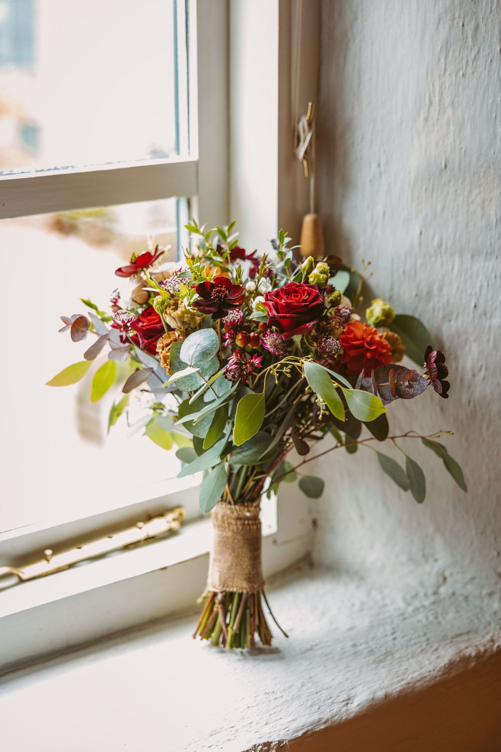A rustic wedding bouquet with red roses and mixed flowers sits on a windowsill in natural light.