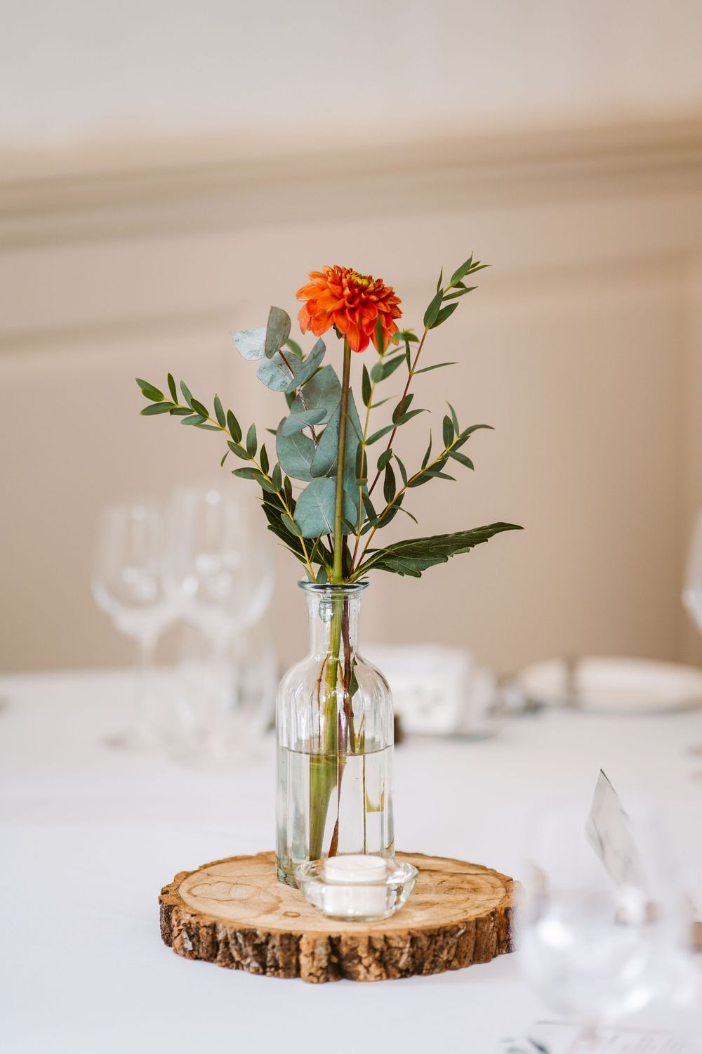 Simple floral centerpiece with orange marigold and eucalyptus in glass bottle on wooden slice.