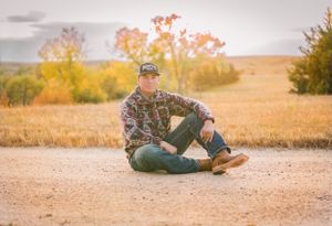 Person in camouflage clothing lying in dry grass field during outdoor photo session.