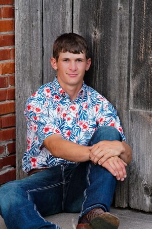 Person in floral shirt and jeans poses casually against weathered wooden barn wall.