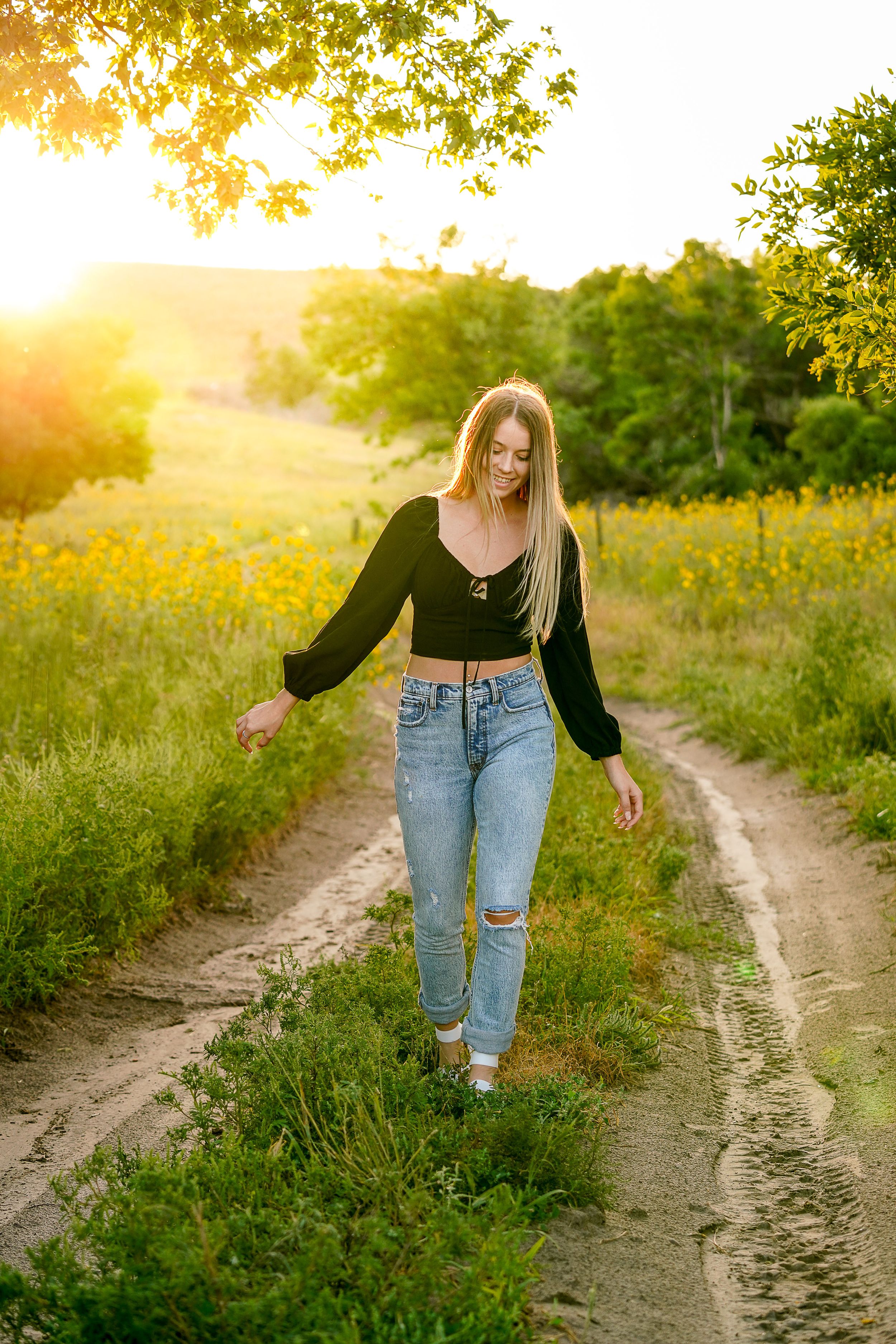 Person walking on dirt path through field at golden hour wearing black top and jeans.