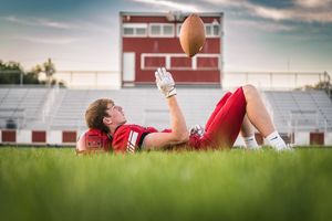 Football player lying in grass field with stadium bleachers visible in background during golden hour sunset.