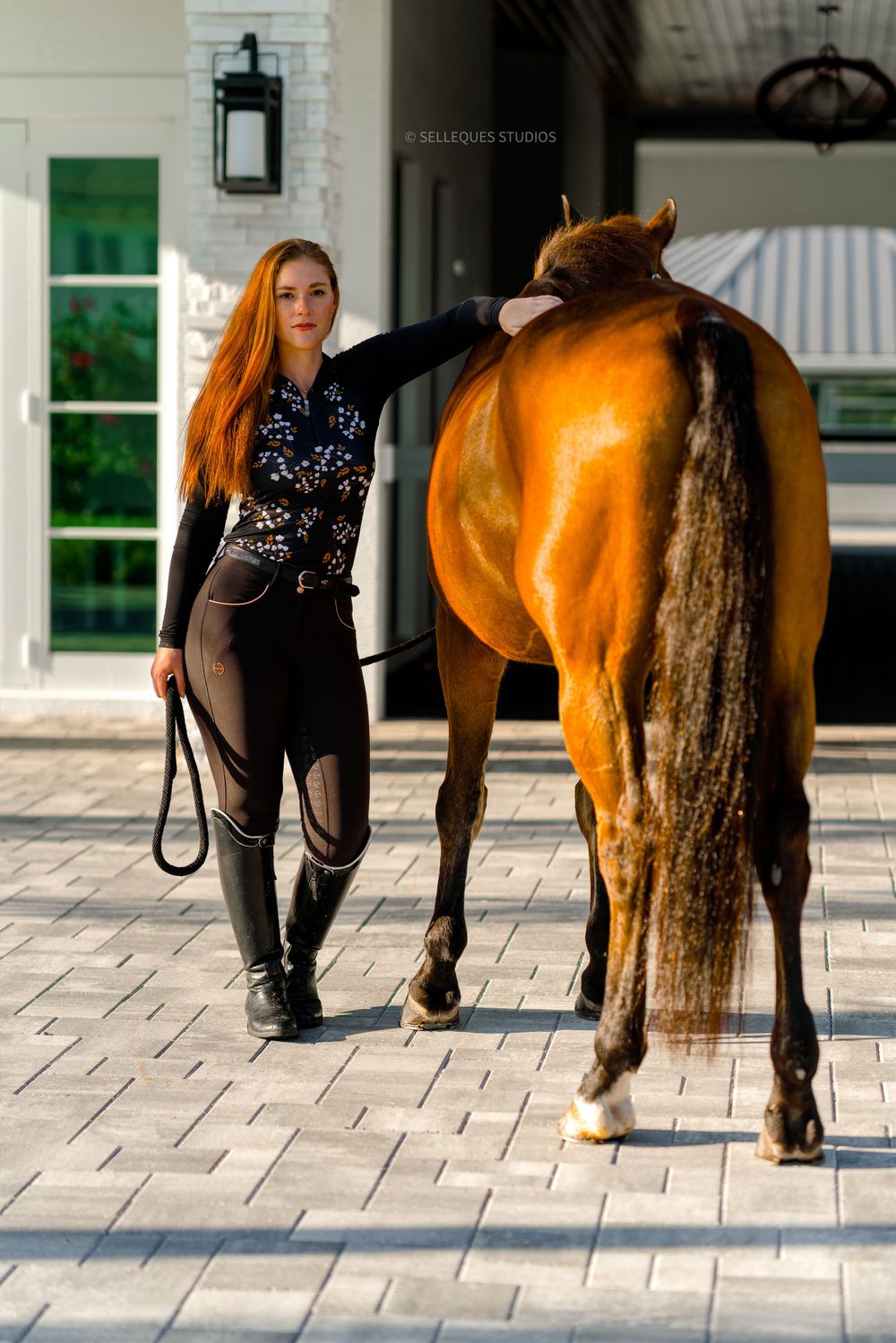 Bay horse and handler walking on decorative stone patio near white building.
