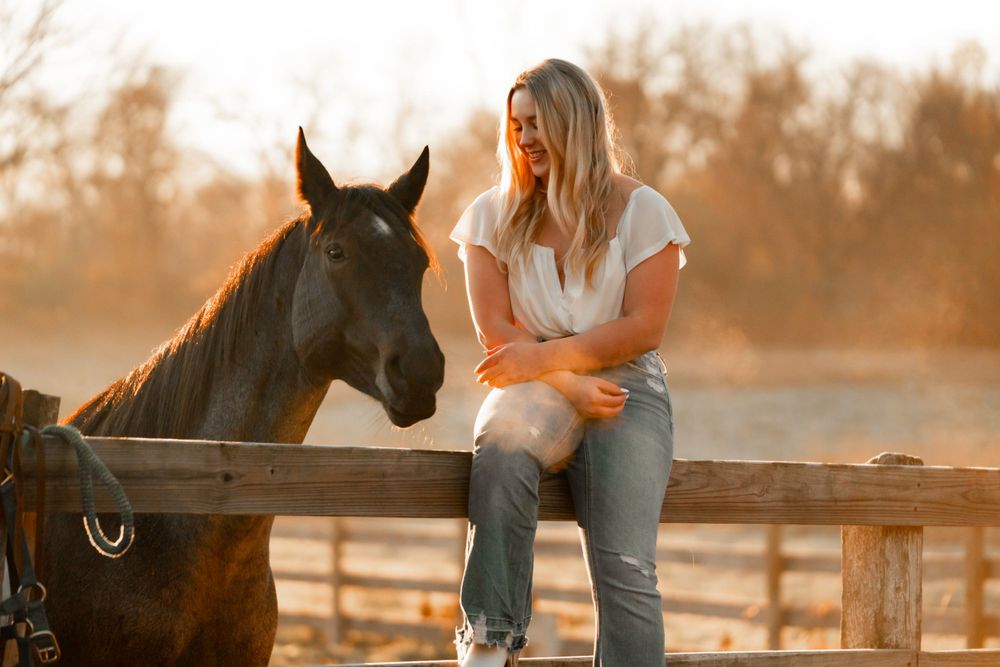 A peaceful sunset moment with a horse and rider sitting on a wooden fence at a rustic farm.