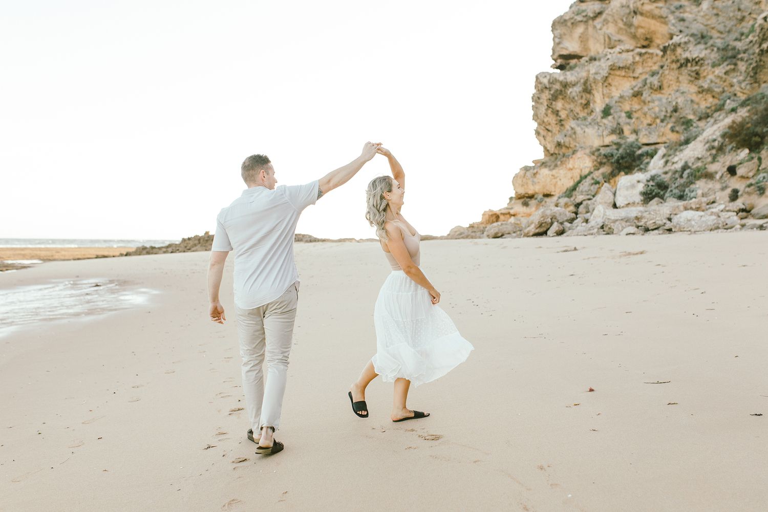 Couple dancing together on a sandy beach with dramatic cliffs in the background.