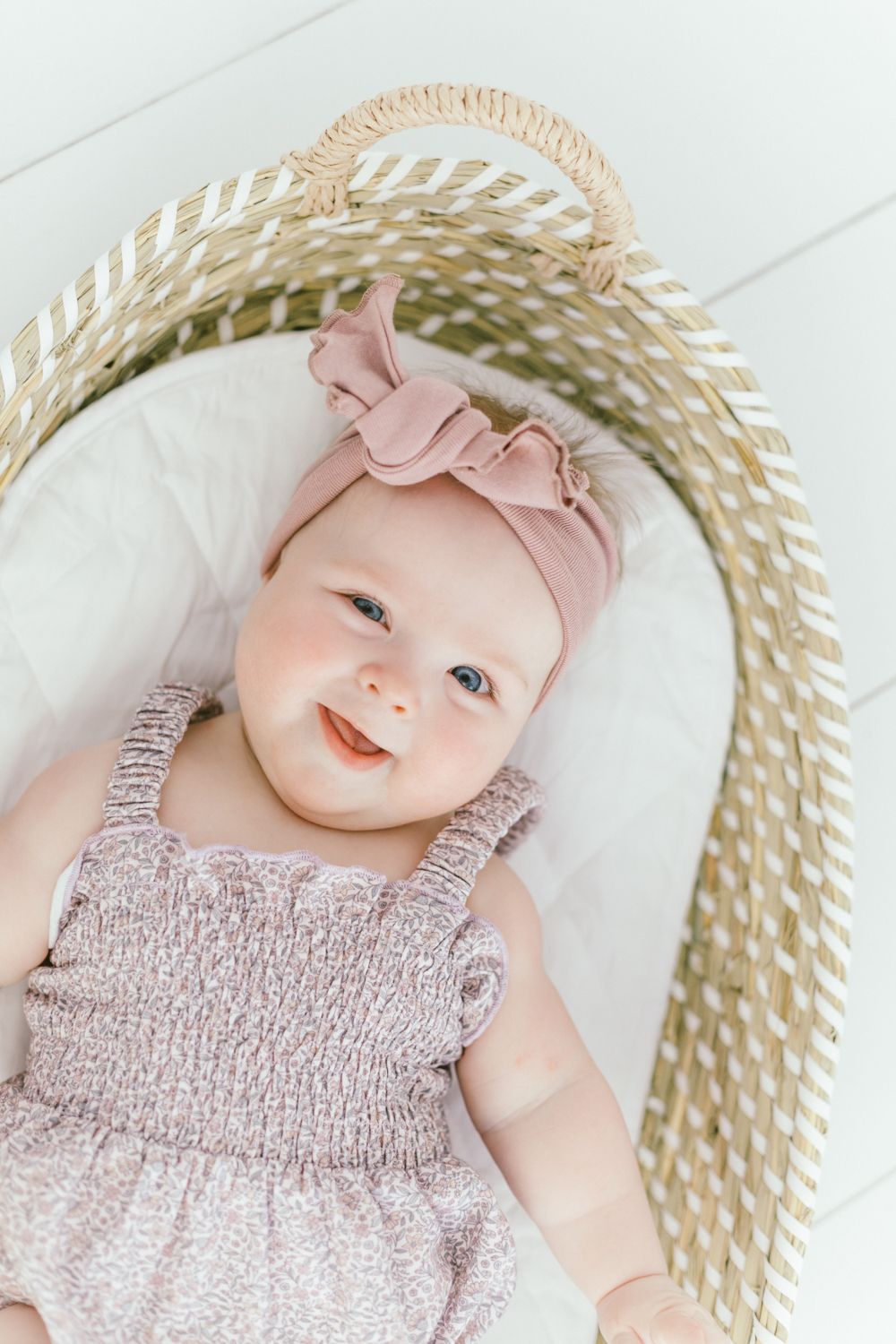 Baby wearing pink ruffled romper and bow headband lies in woven basket with white cushioning.