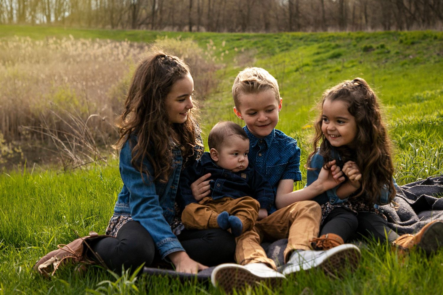 3 kids and their baby brother smiling at each other sitting on a blanket in a grassy field