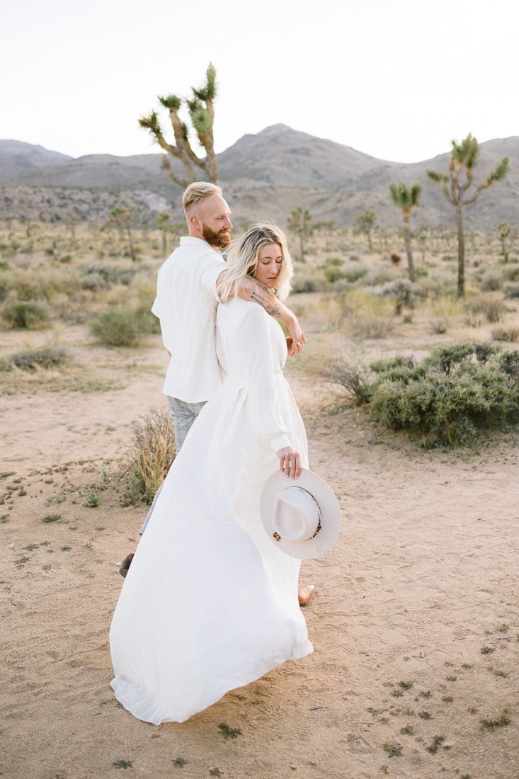 Couple in white clothing posing among desert Joshua trees in a romantic desert setting.