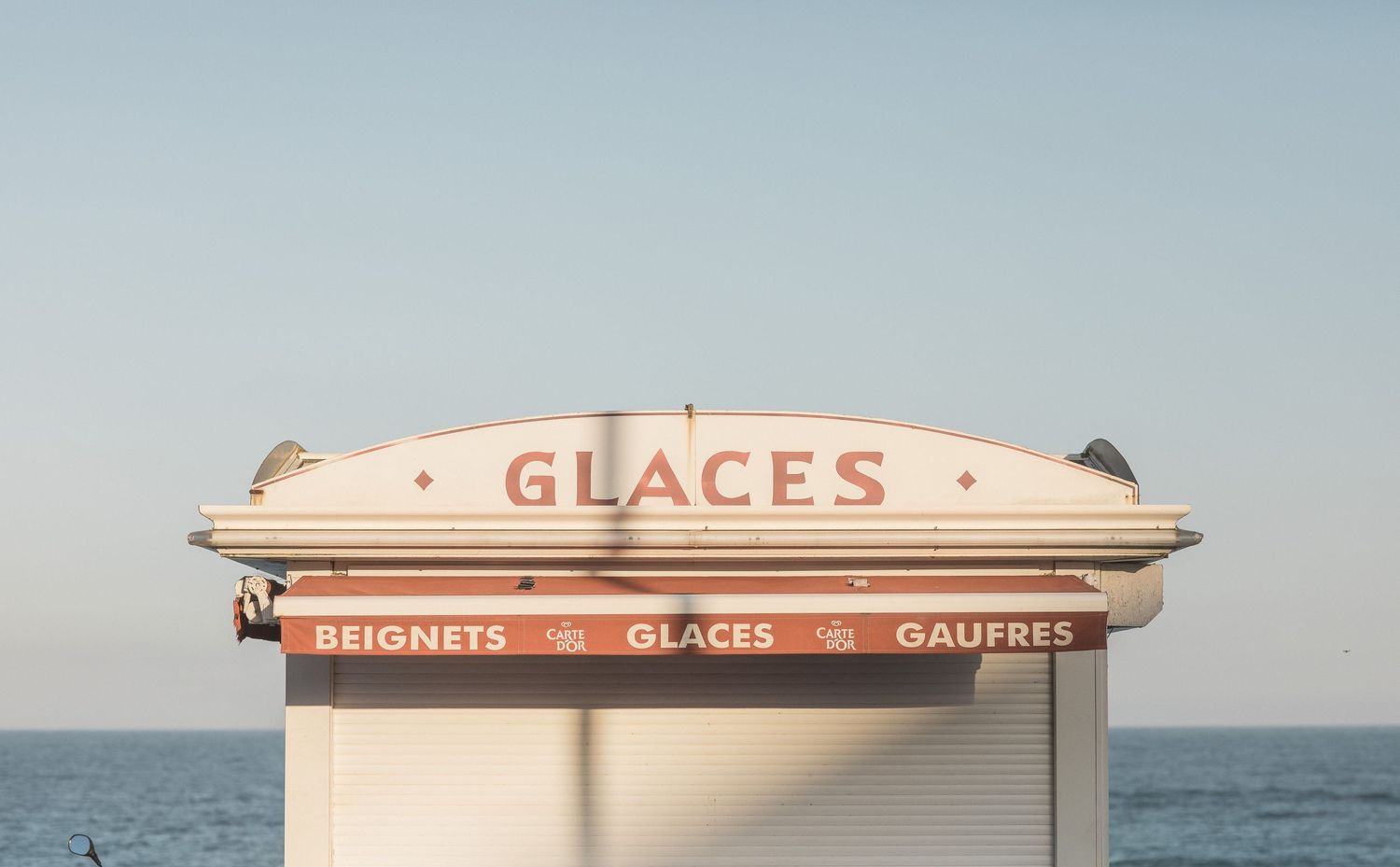 Retro ice cream kiosk sign labeled 'GLACES' against a pale blue sky and ocean backdrop.