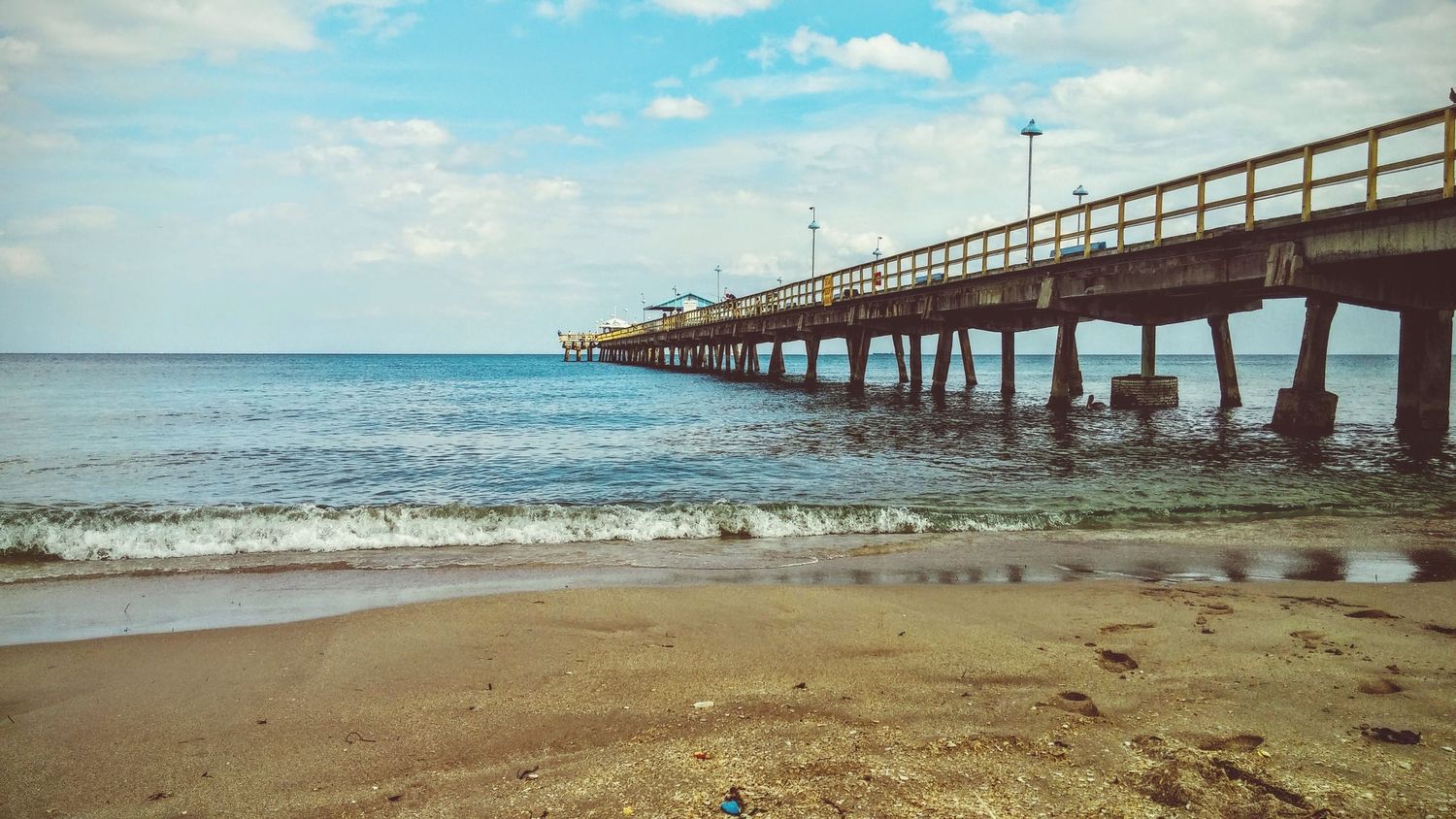 Long wooden fishing pier stretches into calm blue ocean waters on sunny day.