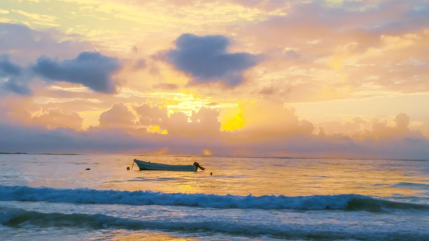 Small boat silhouette on tranquil ocean waters during a golden sunset with clouds reflecting on calm waves.