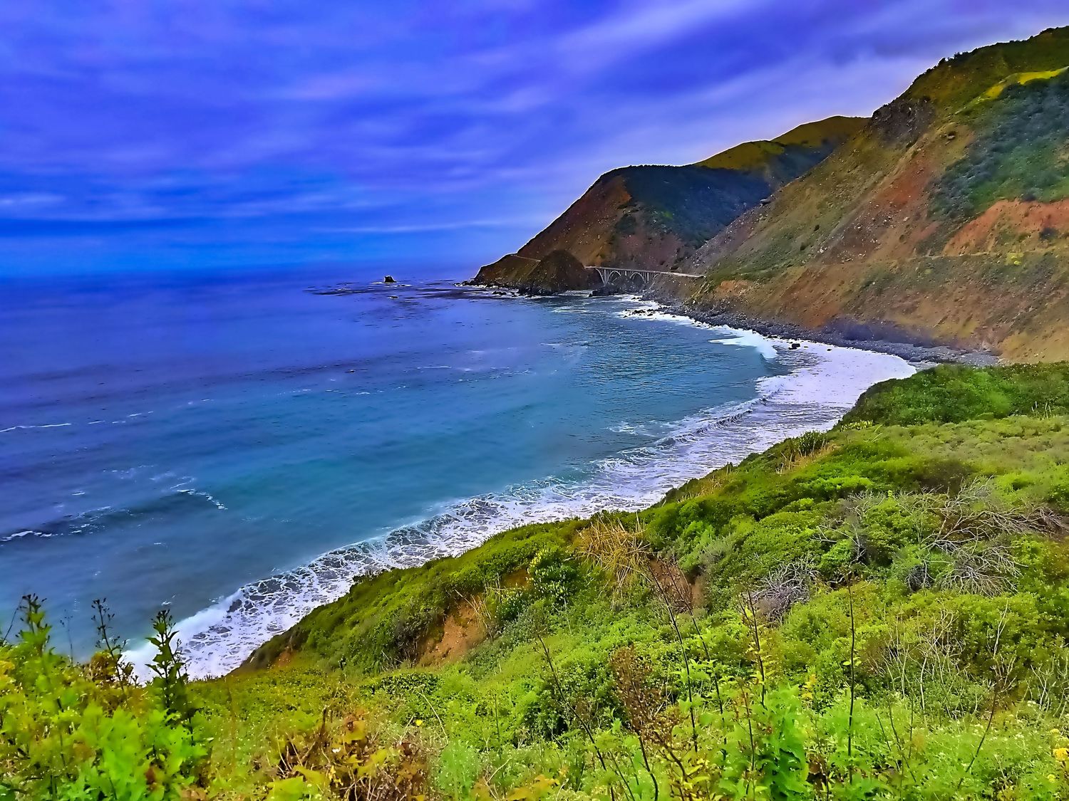 Dramatic coastal landscape with blue ocean waves crashing against green cliffs under vibrant sky.