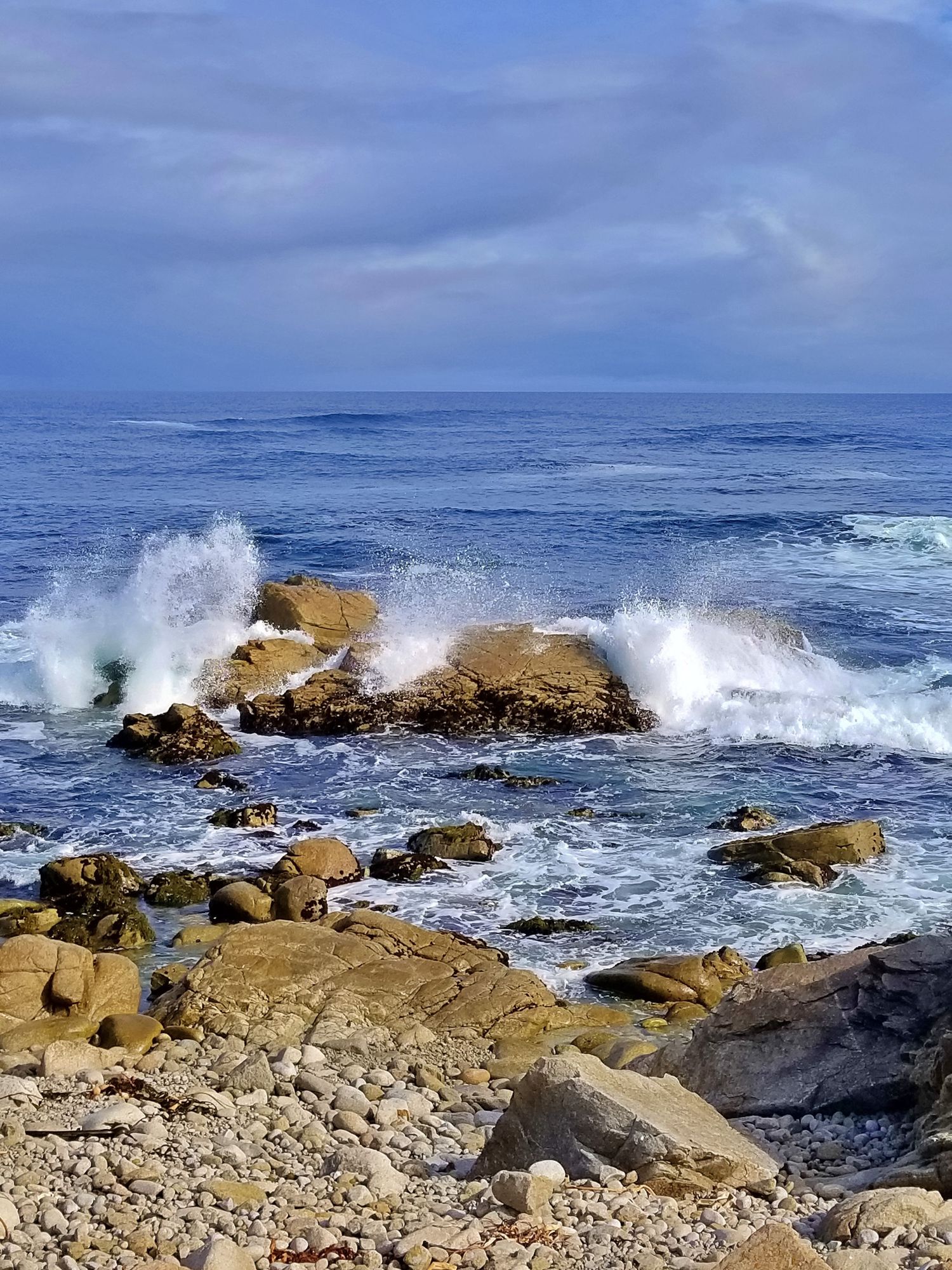 Ocean waves crash against rocky outcrop on rugged coastline under cloudy sky.