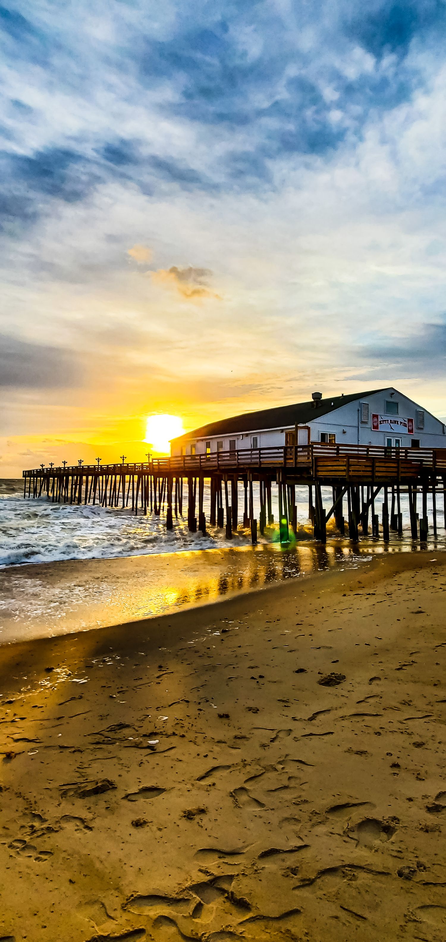 Wooden pier extends over ocean at sunset with golden light reflecting on sandy beach and dramatic clouds above.