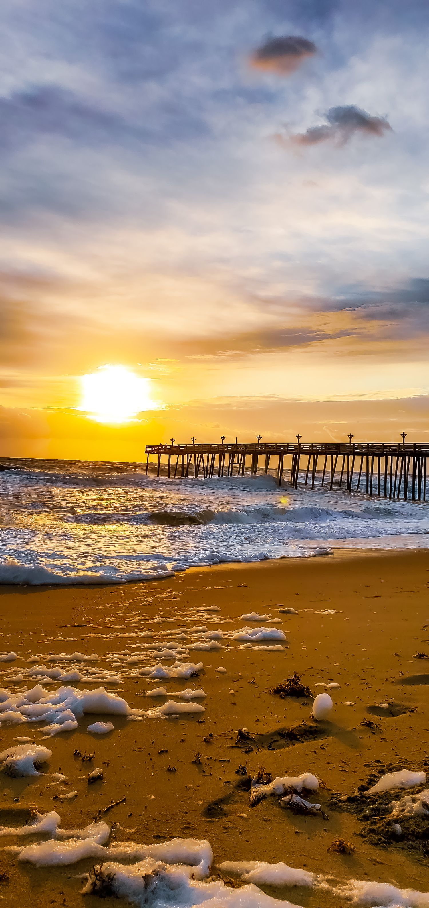 Dramatic sunset over beach pier with sea foam and waves washing onto golden sand.