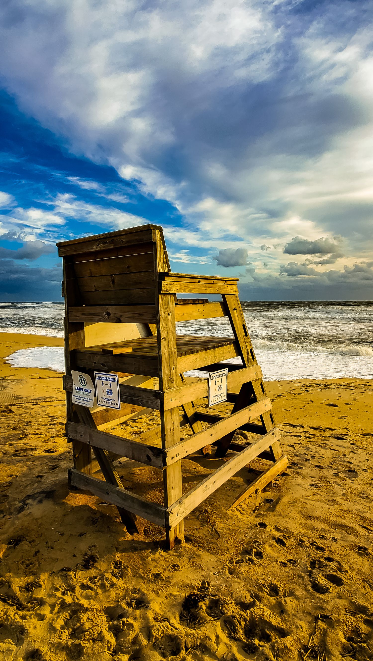 Tall wooden lifeguard tower stands on golden sand beach against cloudy blue sky and rolling waves.
