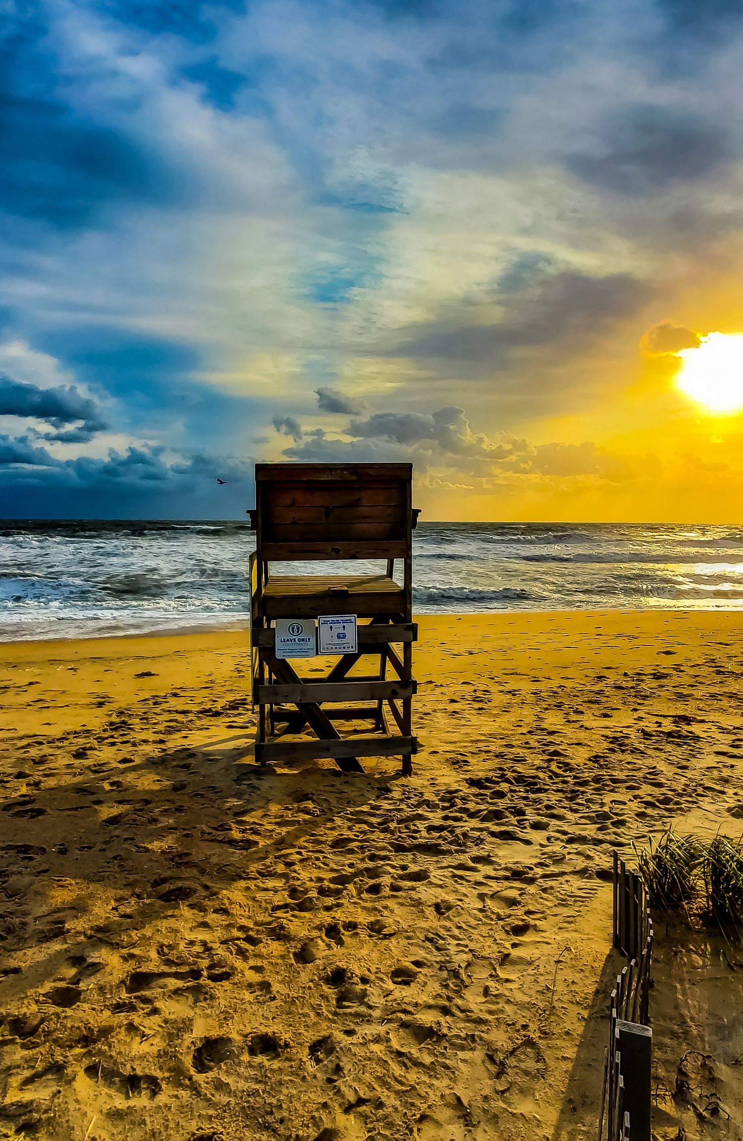 A wooden lifeguard chair sits empty on a beach during a dramatic sunset with stormy clouds and crashing waves.