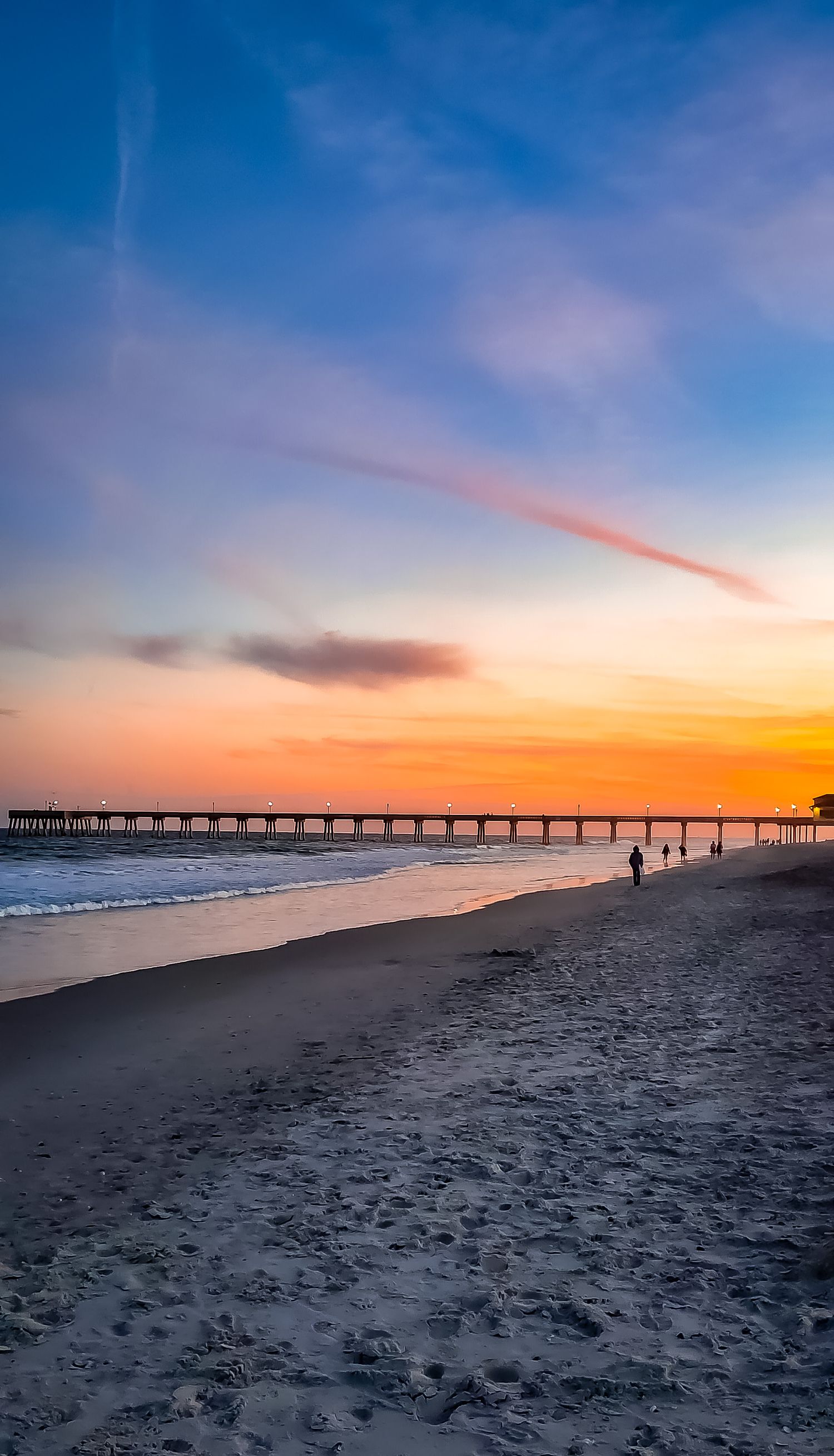 Long wooden pier stretching into ocean at sunset with beach in foreground.