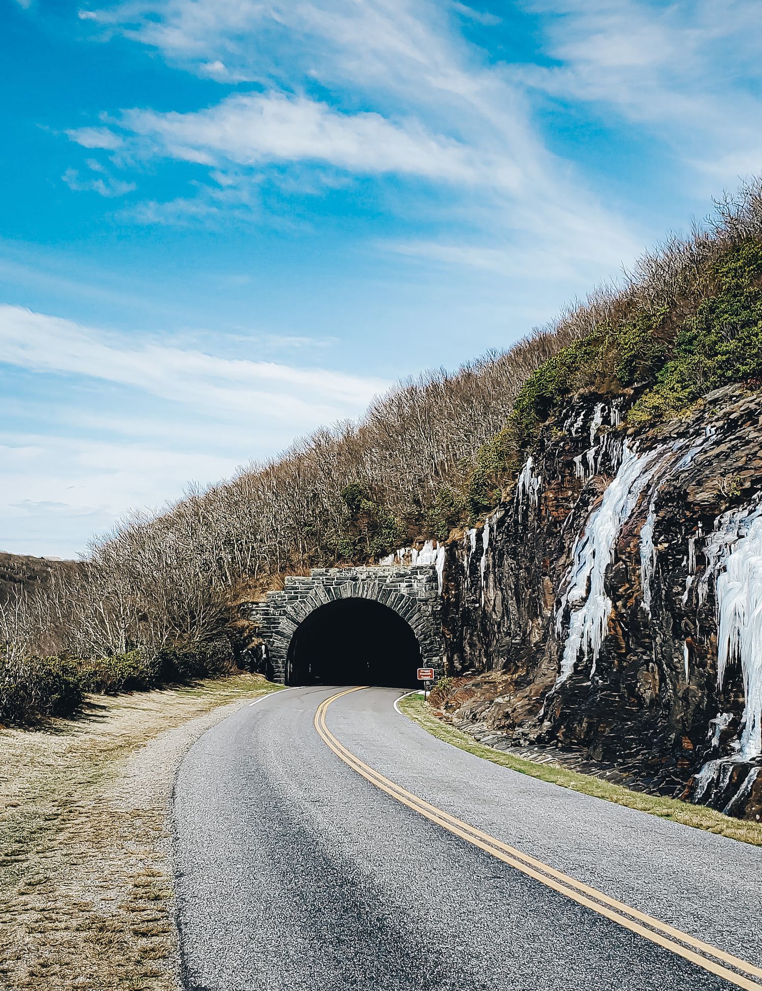 Winding mountain road leads through a rocky tunnel with icicles forming on the cliff face.