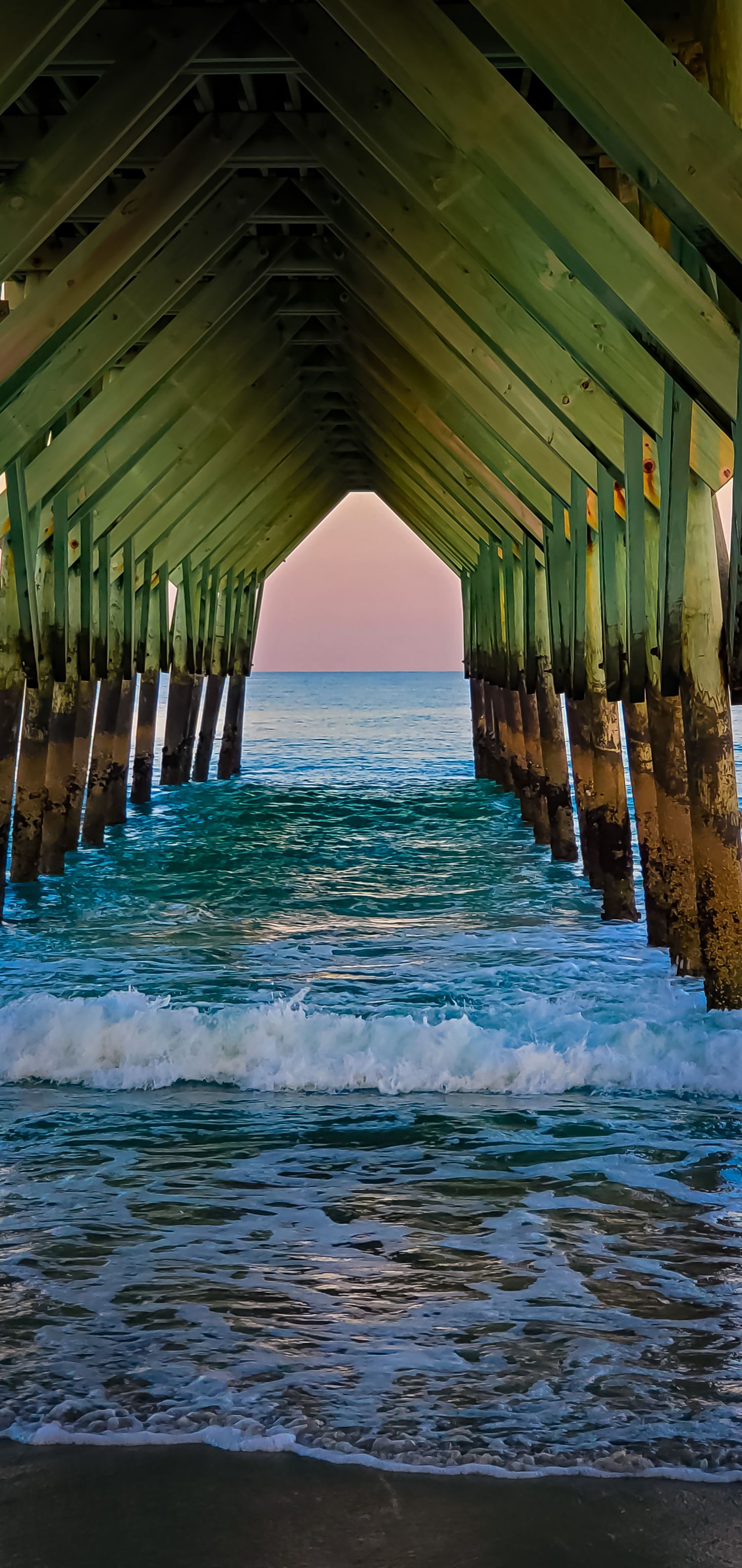 Ocean waves crash beneath a pier creating a symmetrical tunnel view with pink sunset sky in the background.