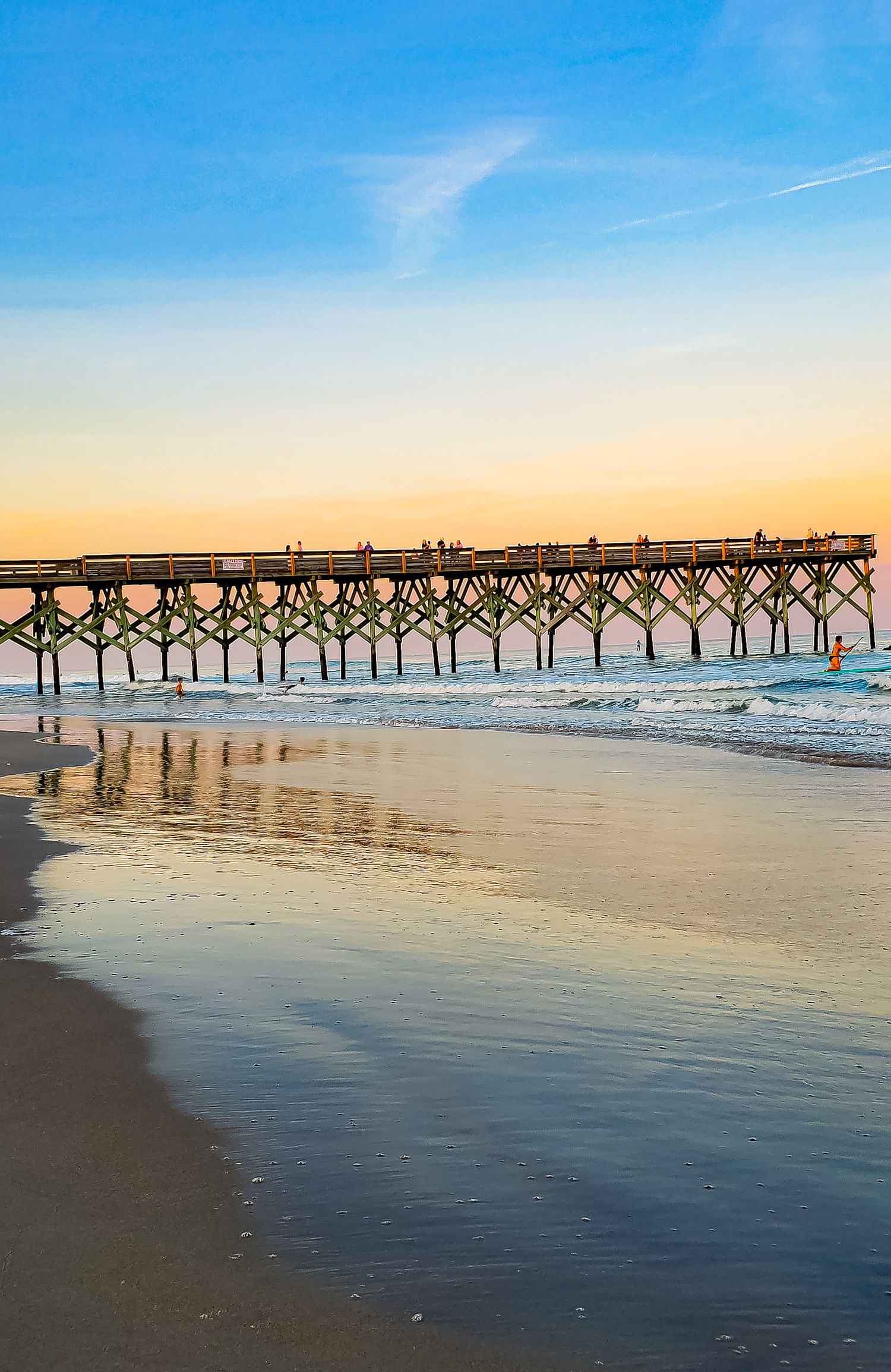 Wooden fishing pier at sunrise with calm ocean waters and reflections on wet sand.