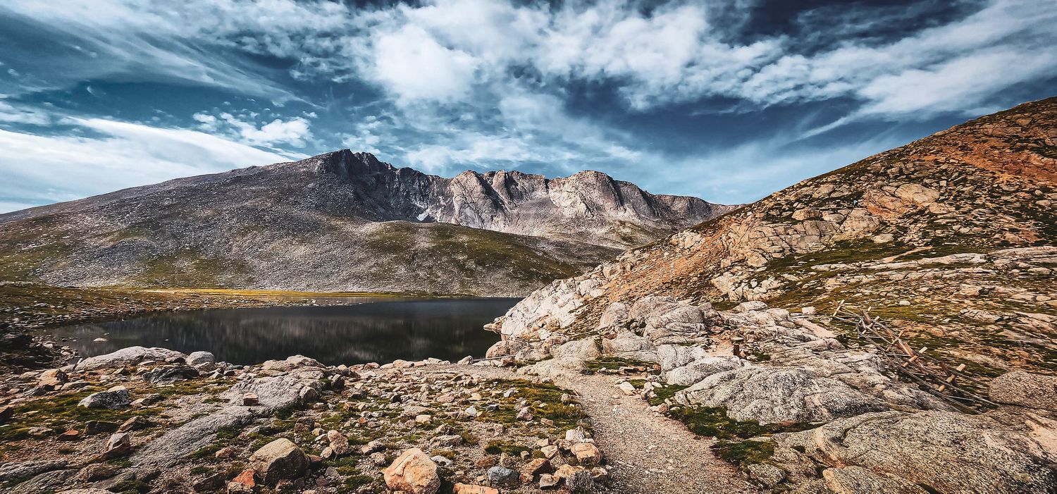 Dramatic rocky mountain landscape with winding trail leading to distant peaks under cloudy skies.