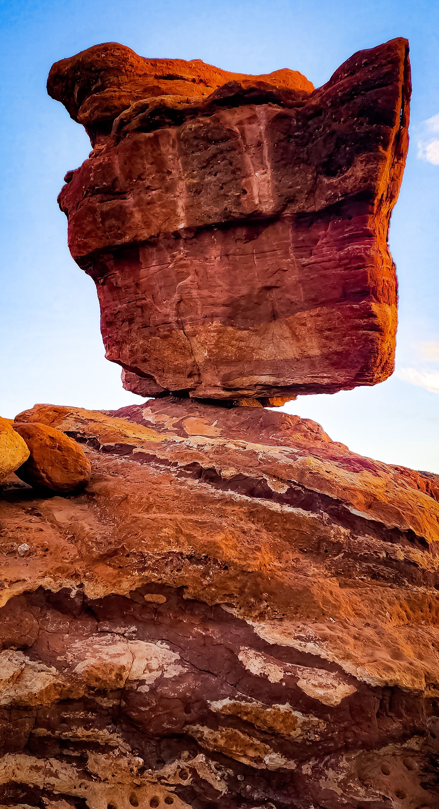 Balanced rock formation stands precariously atop layered red sandstone cliffs in the desert.