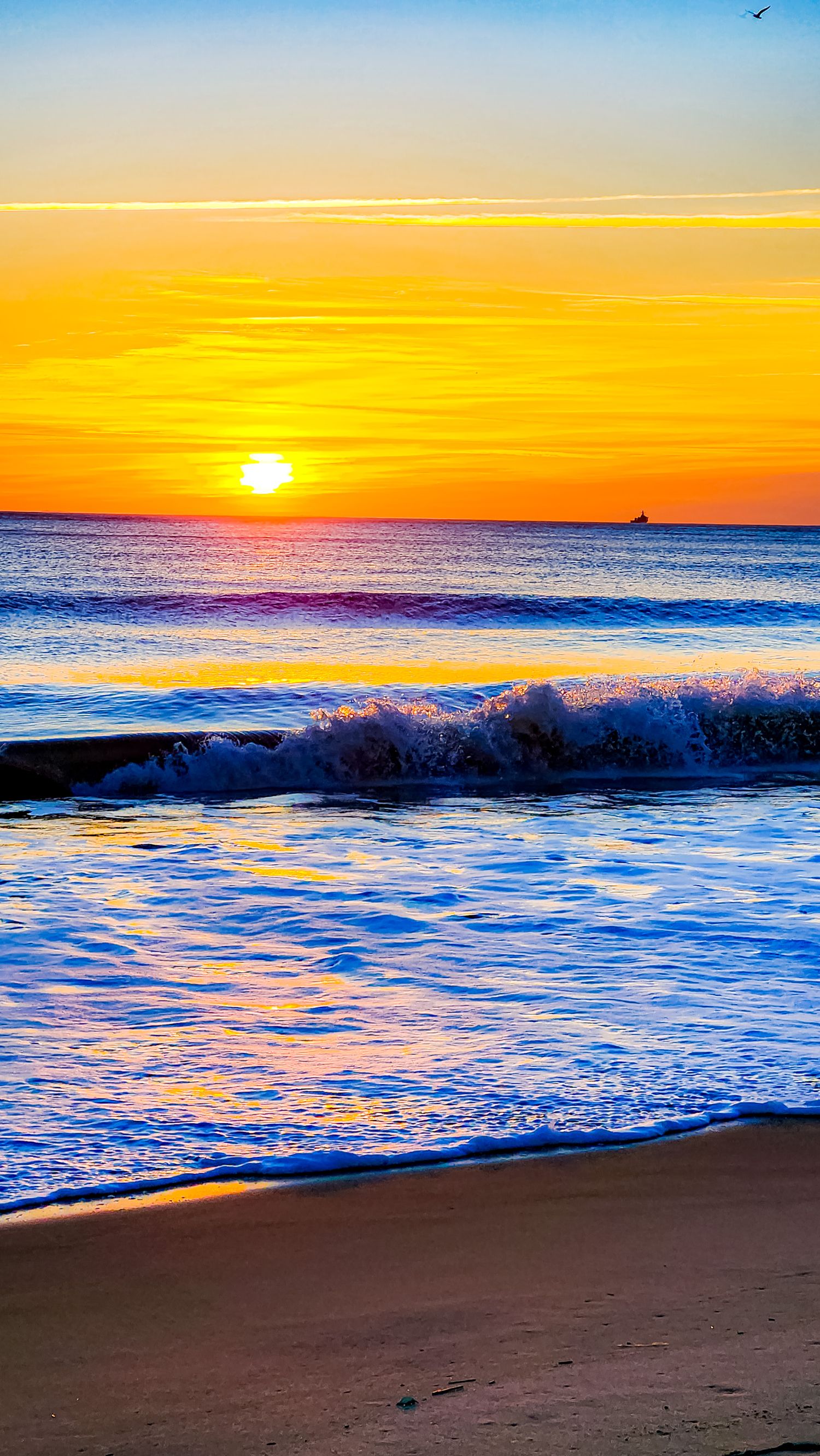 Dramatic ocean waves crash on beach during golden sunset with brilliant orange sky reflection on water.