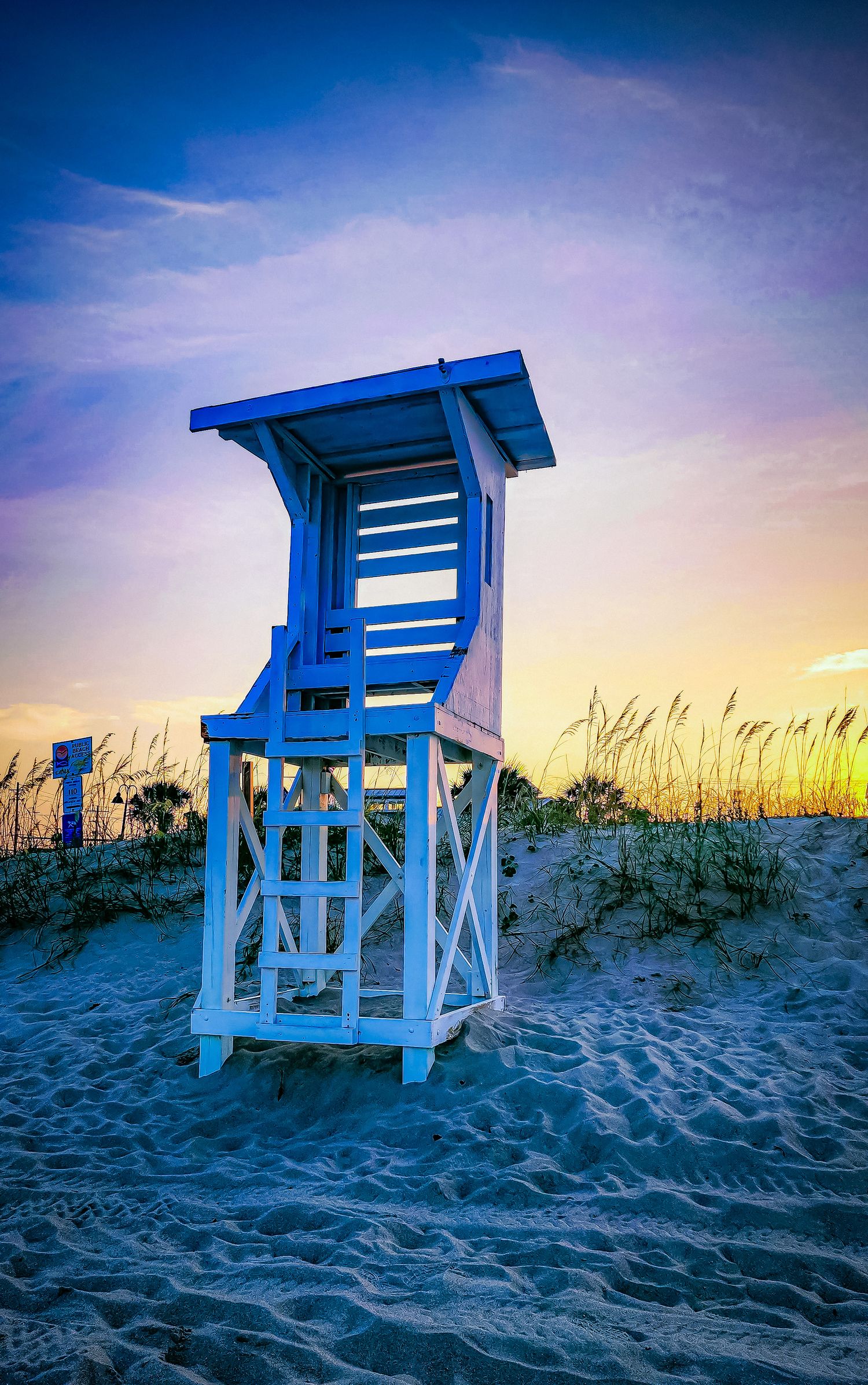 Blue and white lifeguard tower on beach dunes against a purple sunset sky.
