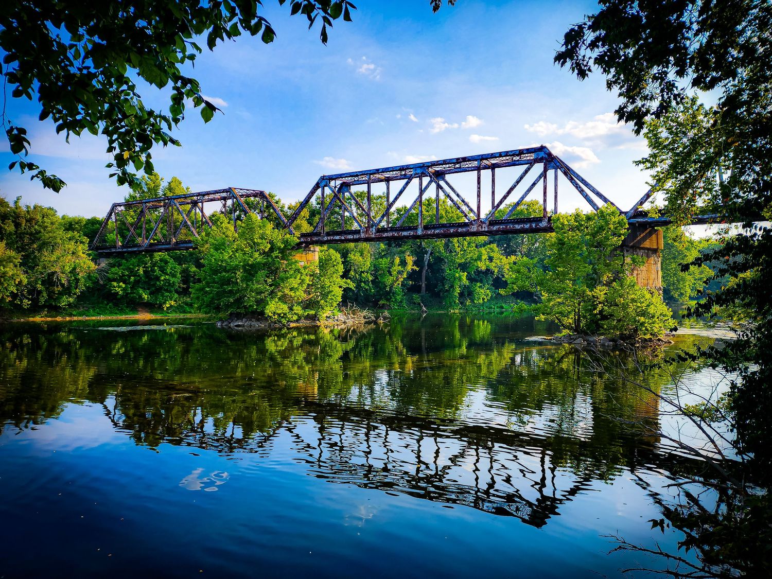 Steel railroad bridge spans across calm river with perfect reflection in summer.
