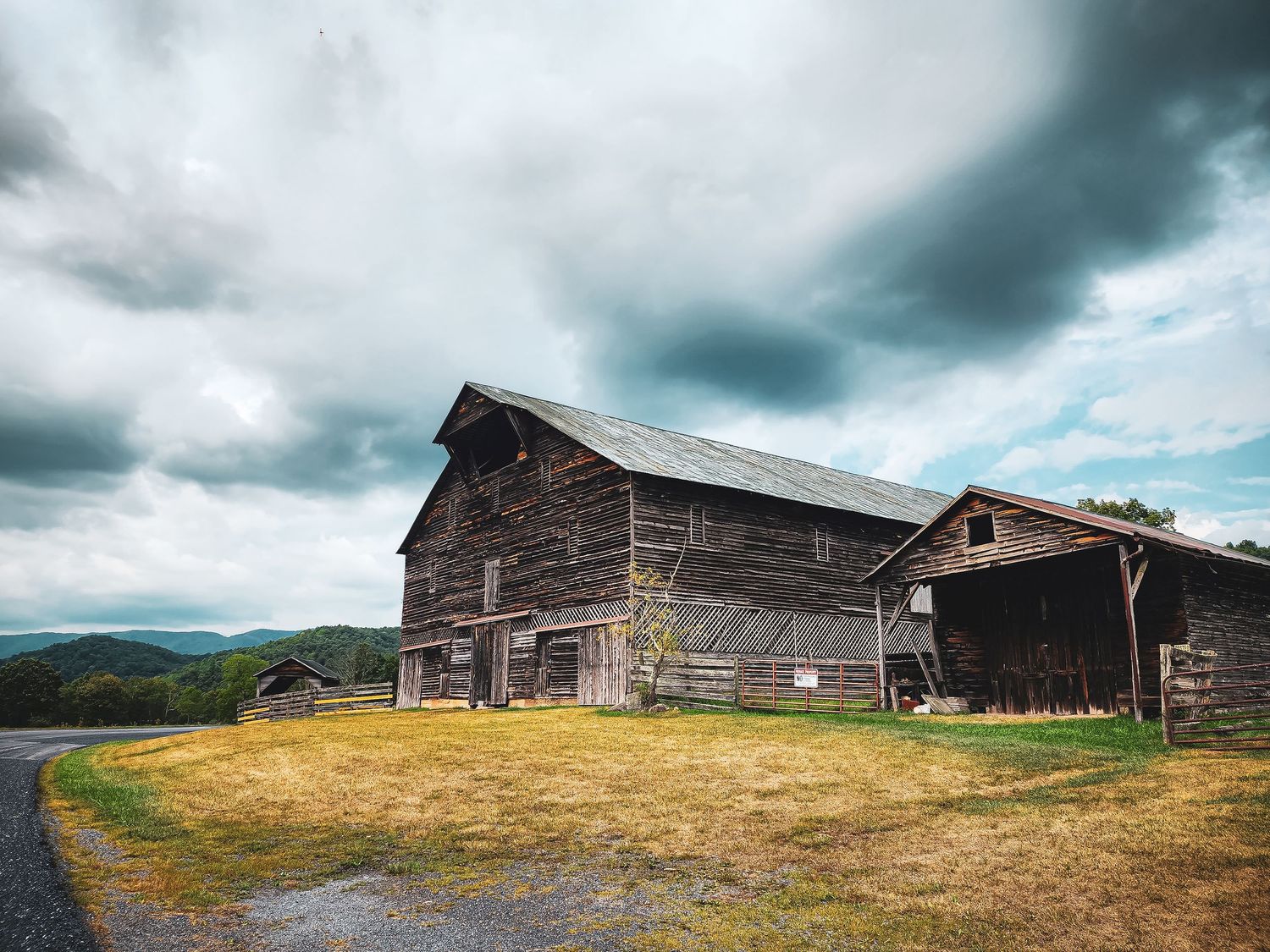 Weathered wooden barn sits atop a grassy hill under dramatic stormy skies in rural countryside.
