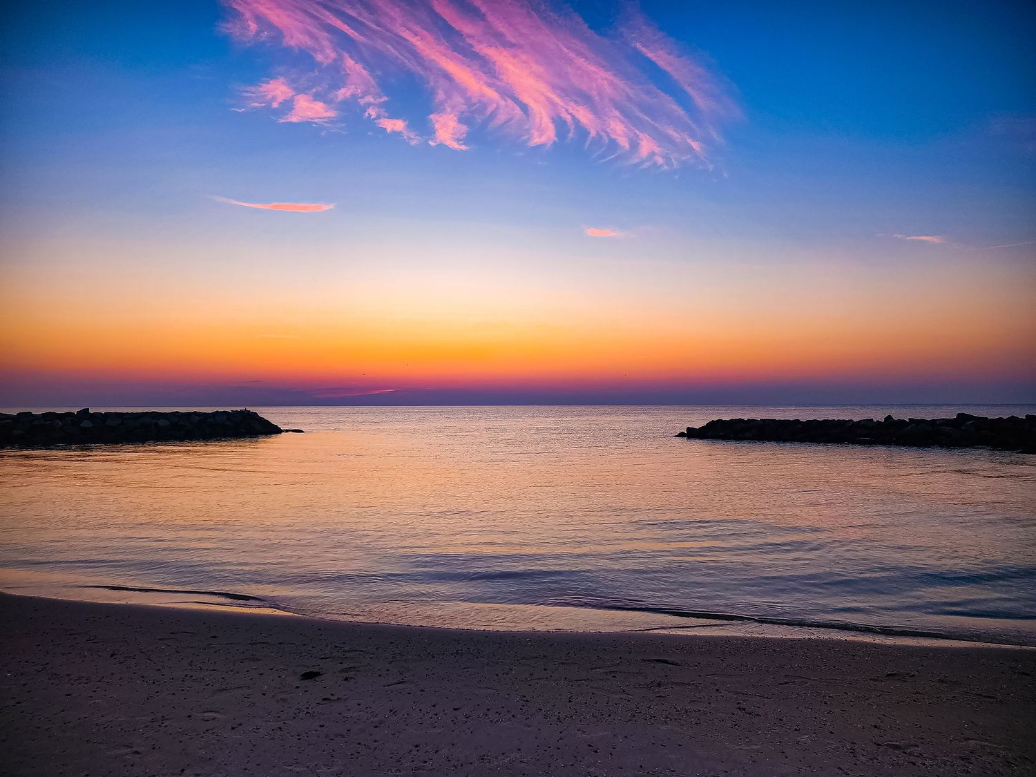 Pink and orange sunset illuminates the calm waters between two jetties at a peaceful beach.
