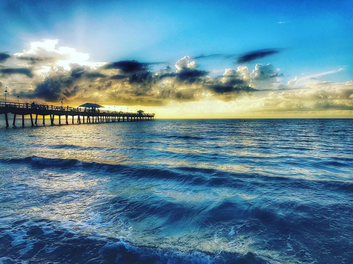 Dramatic sunset over ocean pier with vibrant blue waters and golden clouds.