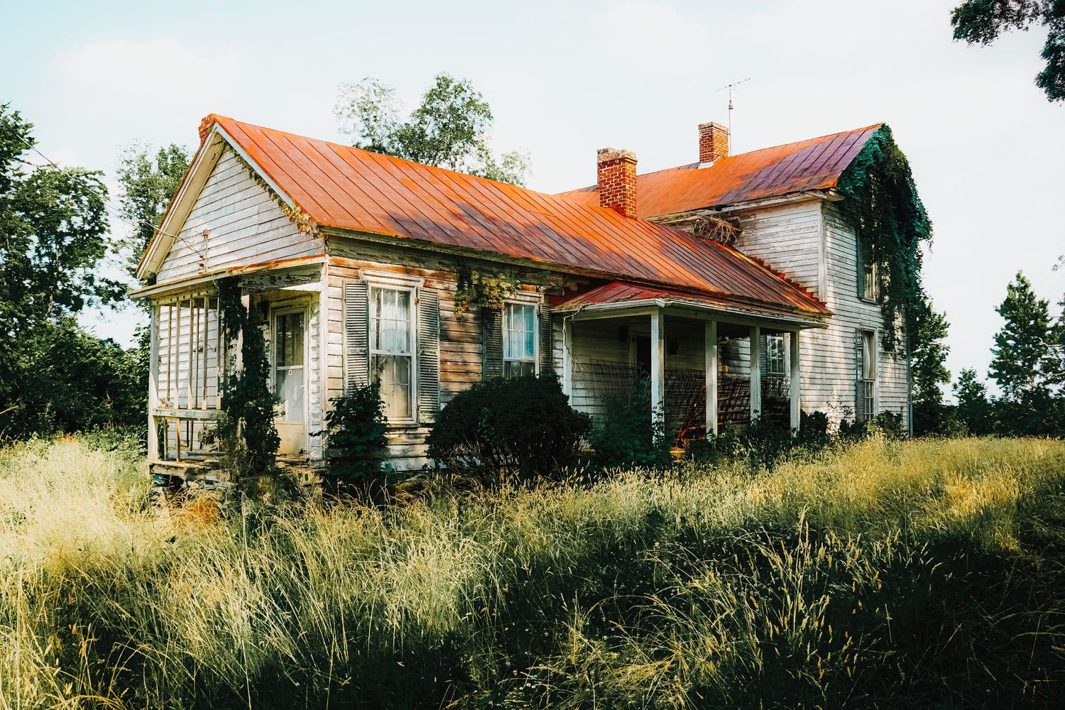 Abandoned rural farmhouse with rusty orange roof and weathered white siding stands among tall grass.