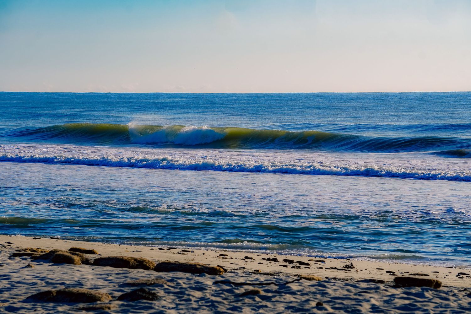Rolling ocean waves with green tint breaking on sandy beach under blue sky.