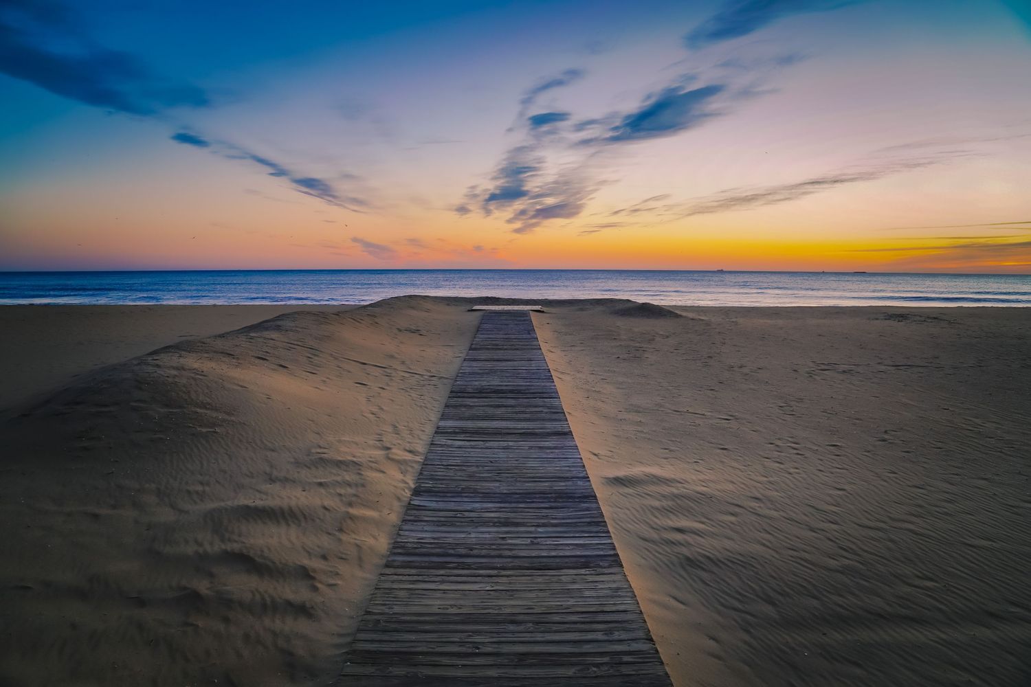 Wooden boardwalk leads to beach horizon during colorful dusk with orange and blue sky.