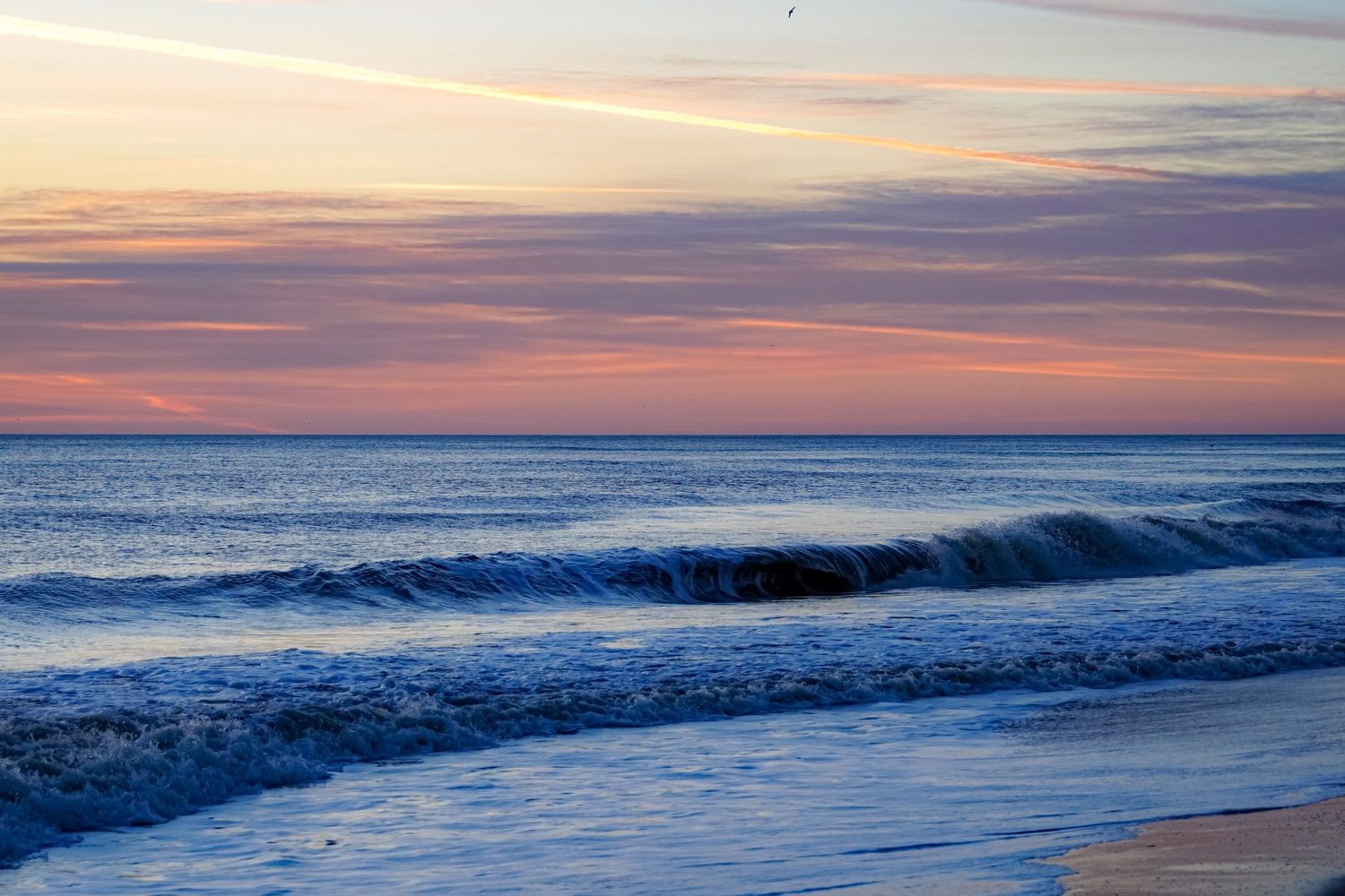 Gentle waves break along shoreline during pink and purple sunset over calm ocean.