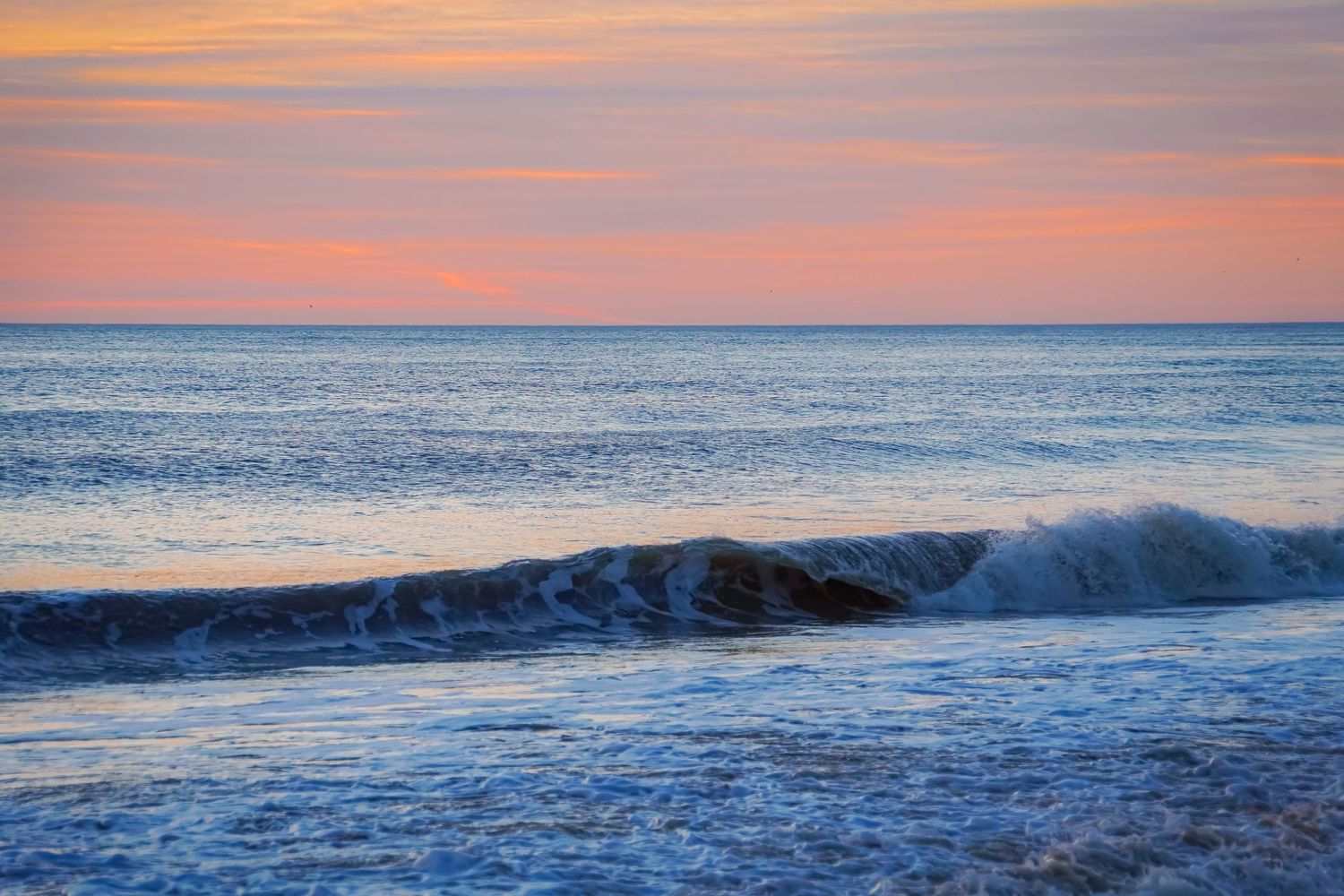 Ocean wave curls and breaks during soft pink sunset with pastel clouds in background.
