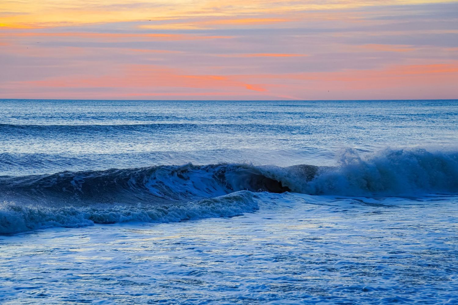 Rolling ocean waves under pink and orange sunset sky with smooth water surface.