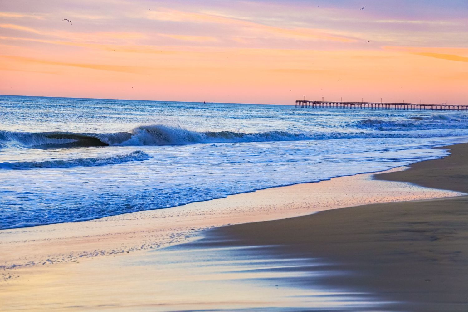 Pastel sunrise illuminates crashing waves and sandy beach near a distant pier.