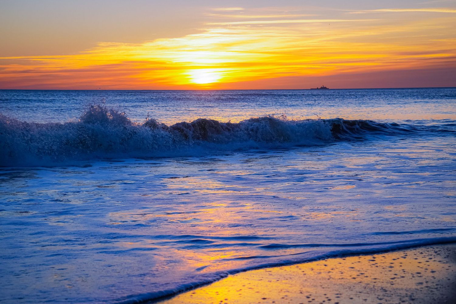 Powerful ocean wave breaks at shore during vivid orange sunset with reflective wet sand.