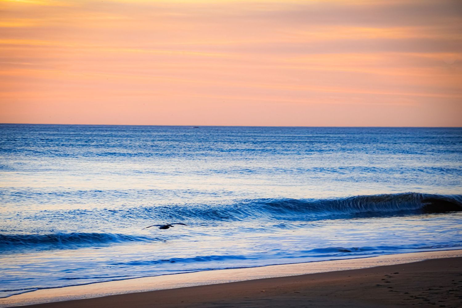 Tranquil ocean waves roll onto shore during a pink and orange sunset with calm waters in the distance.