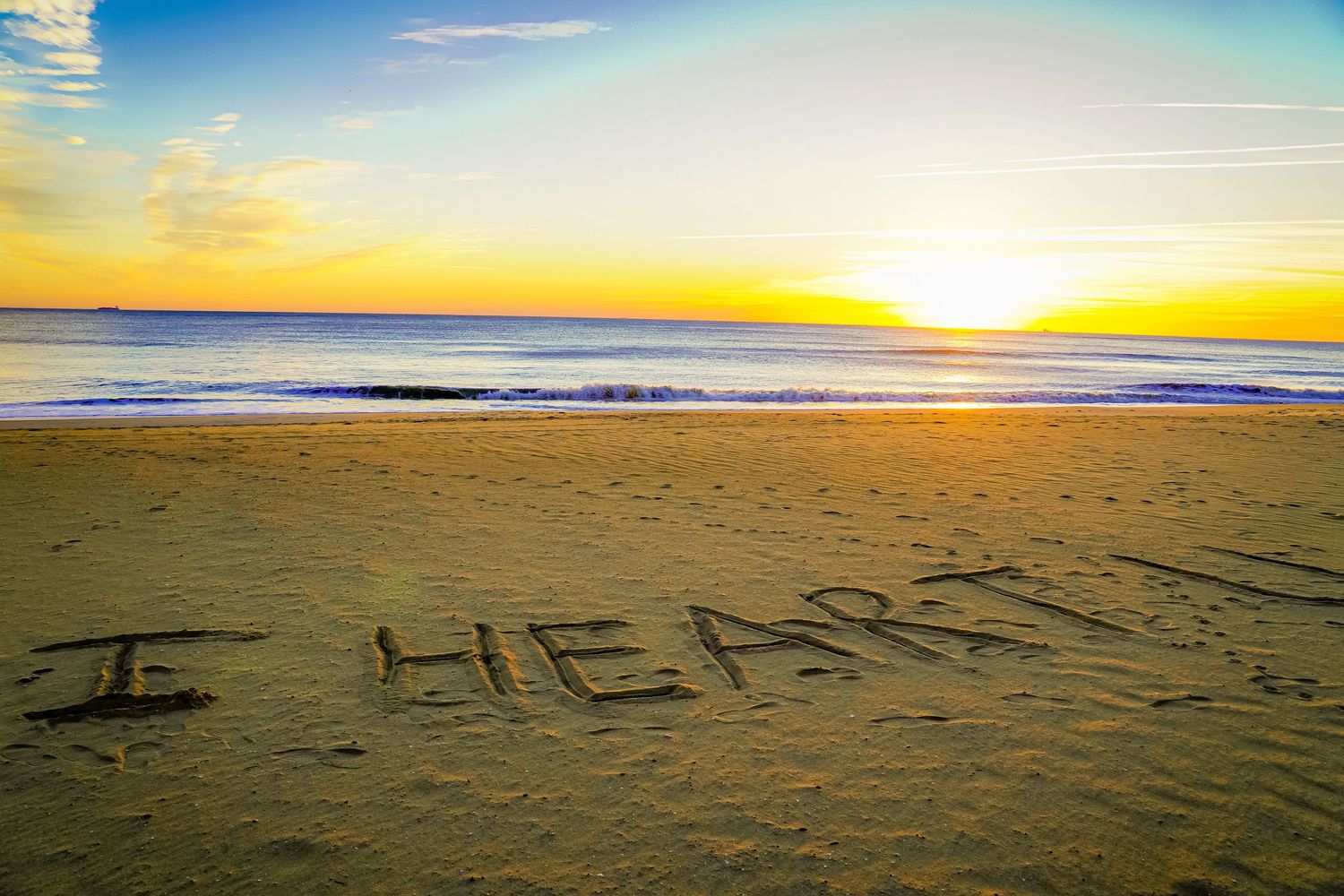 Message reading 'I HEART' written in sand on beach at golden sunset with ocean waves in background.