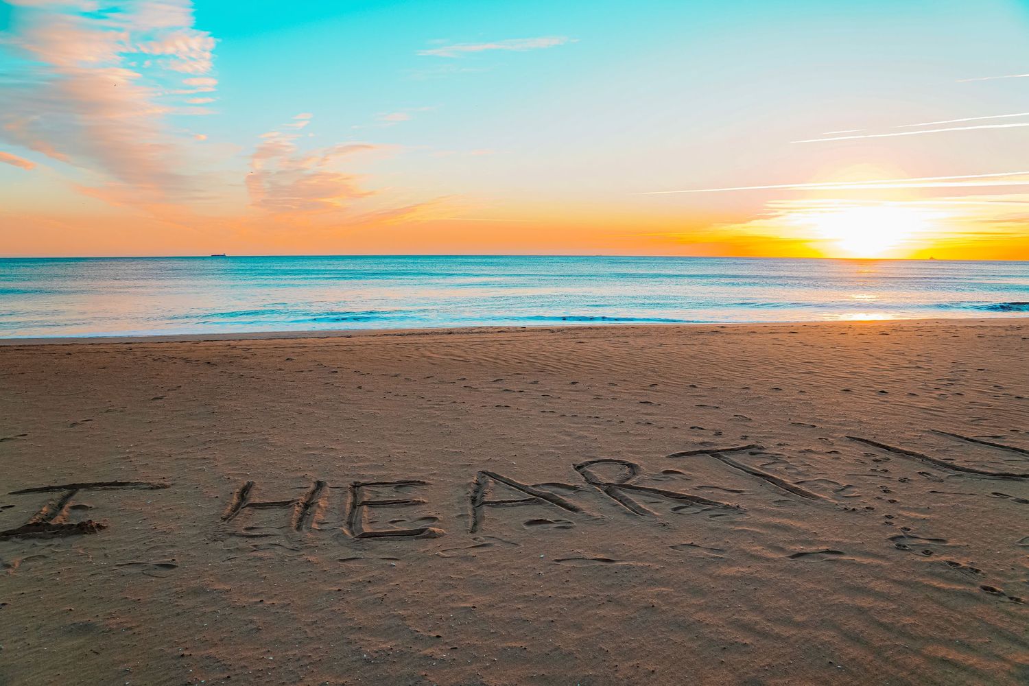 Writing in sand on beach at sunset with turquoise ocean waters and orange sky.