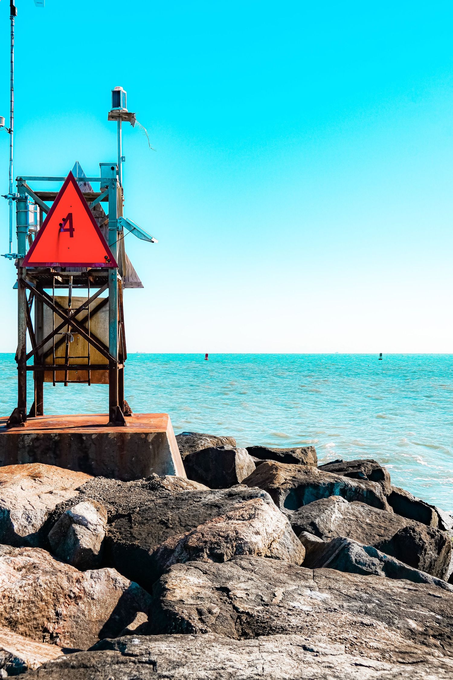 Red triangular navigation marker on wooden platform over rocky coastal outcrop against turquoise sea.